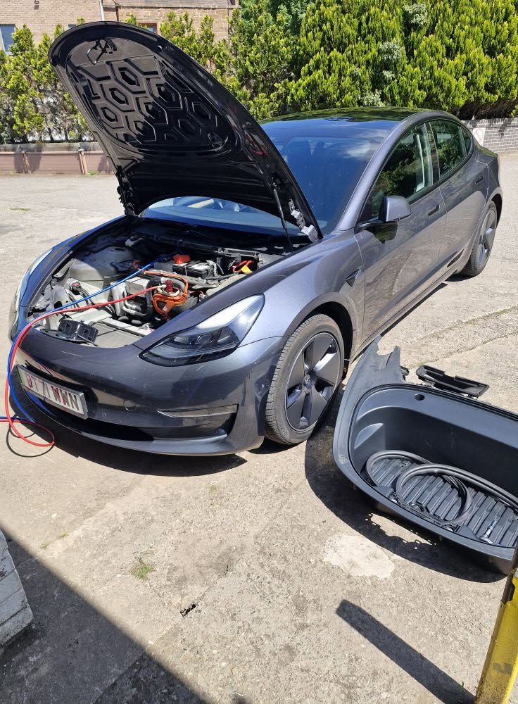 A grey electric saloon with its bonnet up, connected to workshop equipment in the yard