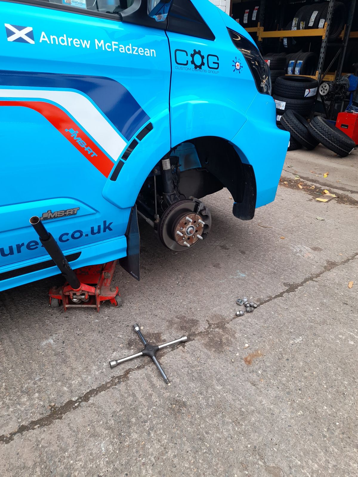 A liveried blue van on a trolley jack with its front wheel off, racks of tyres behind